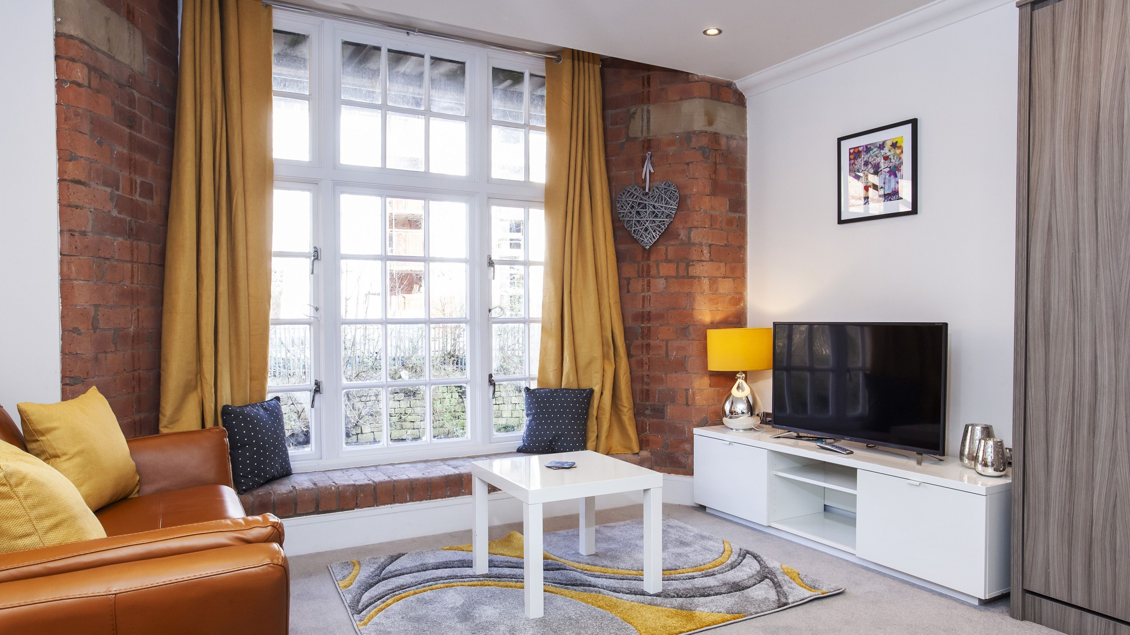 A living room with a leather sofa, a white coffee table, a TV on a white stand, and a large window with curtains.