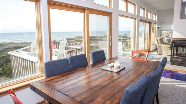 A spacious room with a large wooden table and blue chairs, large windows with ocean views, and a cozy seating area with a fireplace. - Sunny Spot is a unique Oceanfront cottage in Frisco! (FRISCO)