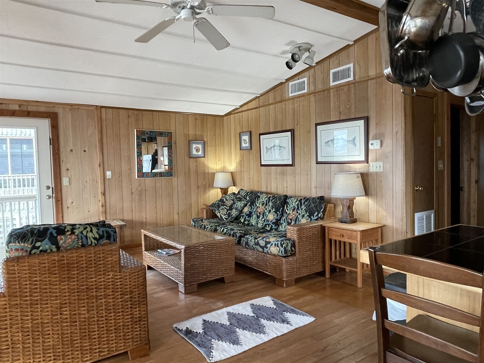 A wooden-paneled room with a ceiling fan, wicker furniture, a glass coffee table, framed pictures on the wall, and a door with a window.