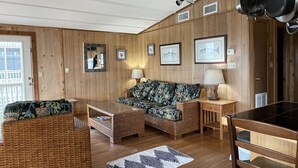 A wooden-paneled room with a ceiling fan, wicker furniture, a glass coffee table, framed pictures on the wall, and a door with a window. - Hatteras Jack Oceanfront Cottage with a great ocean view! (Hatteras)