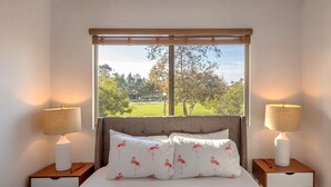 A bedroom with a large window, two bedside tables with lamps, a bed with white bedding and flamingo patterned pillows, and a view of a green landscape.