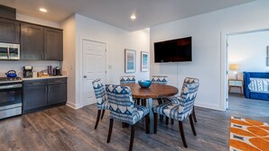 A modern kitchen with dark cabinets, a built-in oven, and a microwave. There is a dining area with a round table and patterned chairs. A flat-screen TV is mounted on the wall above the table.