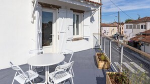 A white building with a tiled roof, a balcony with a table and chairs, and a view of the street.