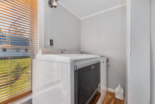 A compact laundry room with a front-loading washer and dryer, a small window with blinds, and a visible water meter.