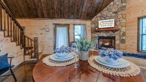 A wooden cabin interior with a stone fireplace, a dining table set with plates and glasses, and a staircase.