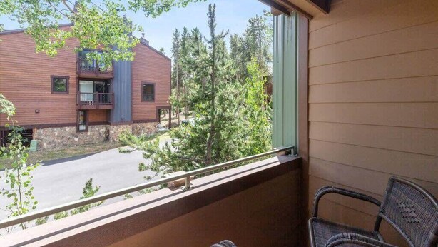 Un balcon avec un fauteuil en osier, une vue sur un immeuble doté d’un balcon, et des arbres en arrière-plan.