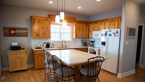 A kitchen with wooden cabinets, a large island with bar stools, a refrigerator, and a window with blinds. - Lake Greenwood, Big Water and Sunset Views (Chappells)