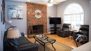 A living room with a brick fireplace, leather sofas, a flat-screen TV, and a large arched window. - Lake Greenwood, Big Water and Sunset Views (Chappells)
