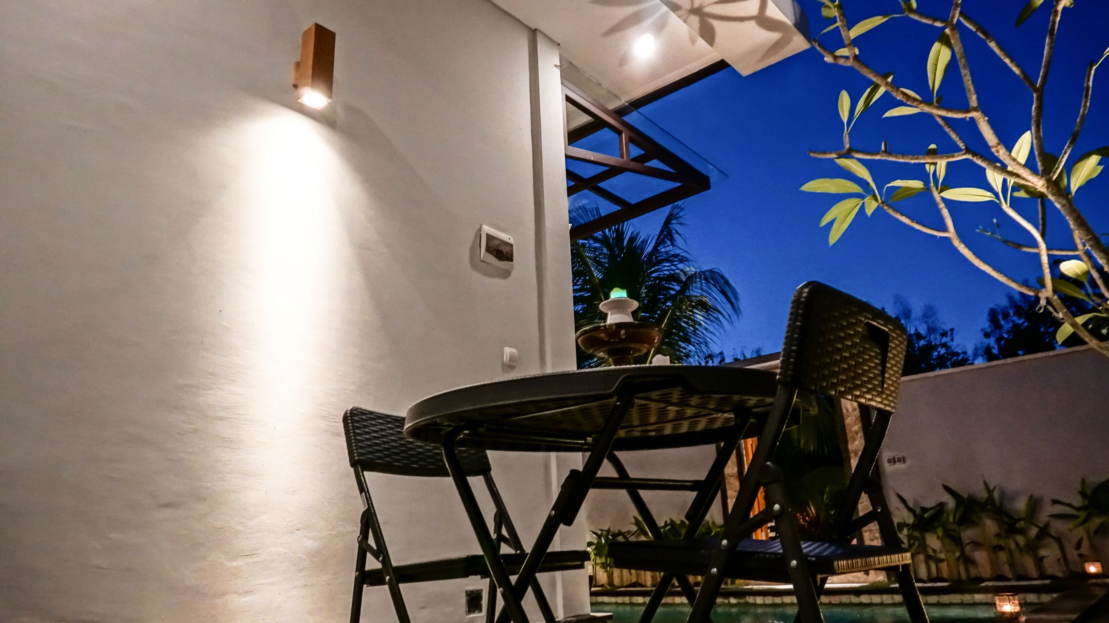 Un espacio al aire libre con mesa y sillas, un aplique en la pared y vistas al cielo y árboles.