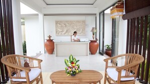 Un espace d’accueil comprenant un comptoir de réception, une réceptionniste, deux chaises en bois, une table agrémentée d’un arrangement floral et de grands vases.