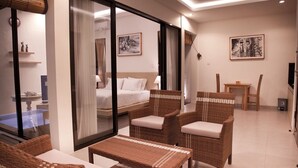 Une chambre d’hôtel moderne comprenant un lit, deux fauteuils en osier, une petite table et une cloison en verre qui sépare la chambre d’un espace piscine.
