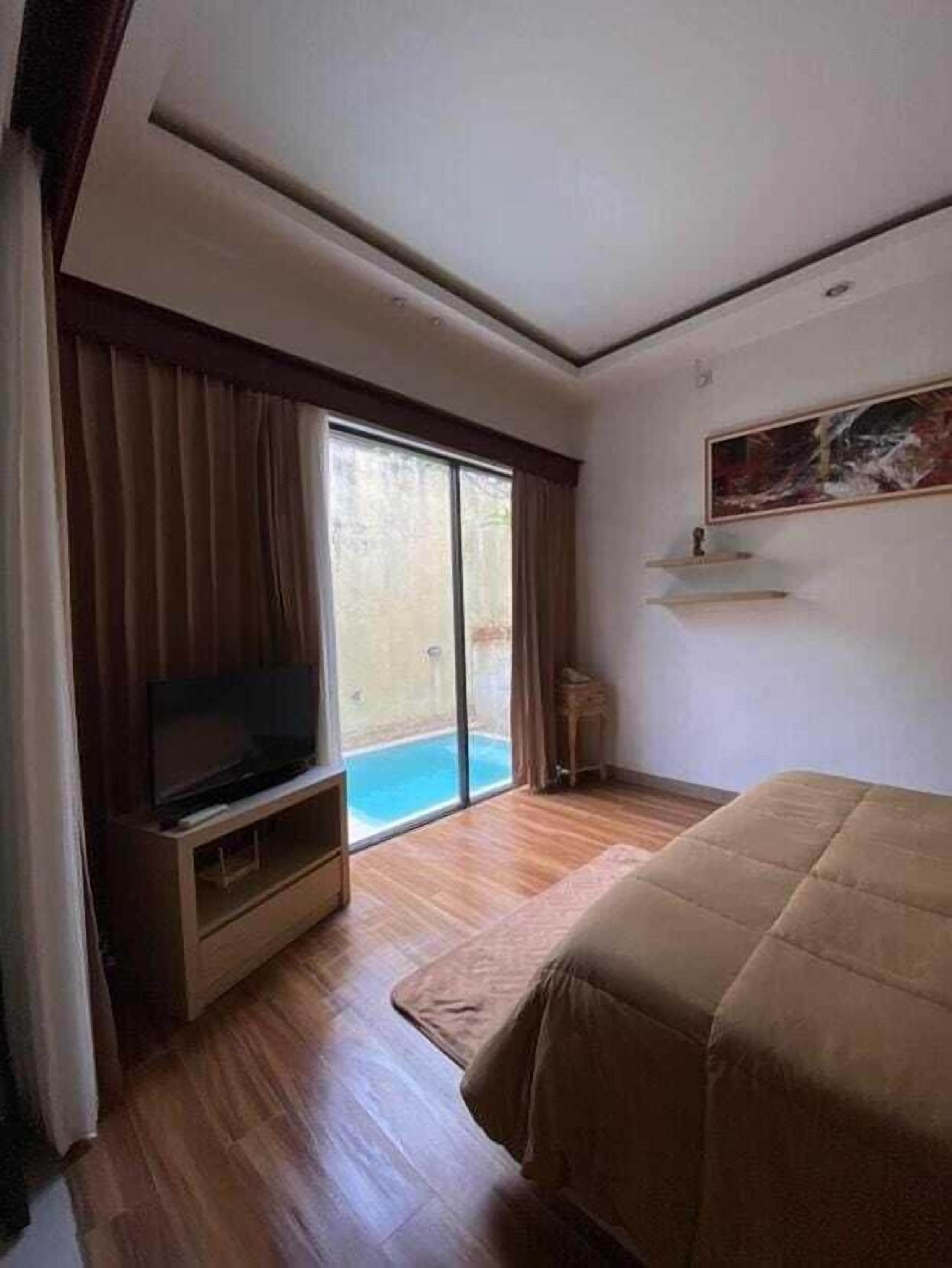A hotel room with a large bed, a television on a wooden stand, a painting on the wall, and a sliding glass door leading to a pool.