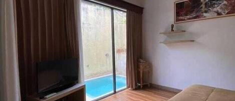 A hotel room with a large bed, a television on a wooden stand, a painting on the wall, and a sliding glass door leading to a pool.