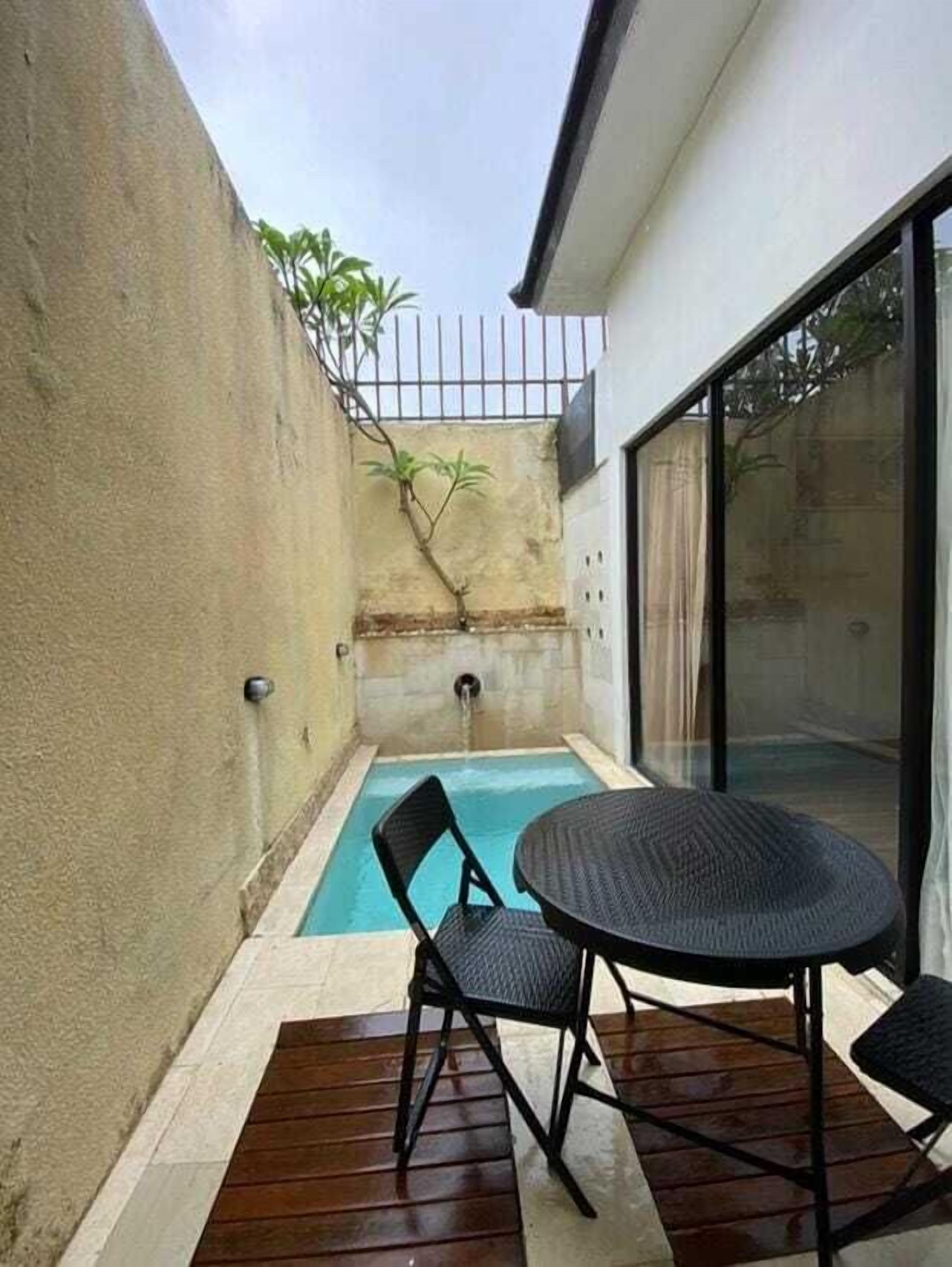 A small outdoor pool area with a single chair and a table, surrounded by a concrete wall and a glass door leading to a covered patio.