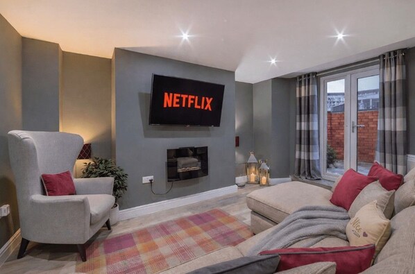 A living room with a flat-screen TV mounted on the wall, a grey armchair, a sofa, and a colorful rug.