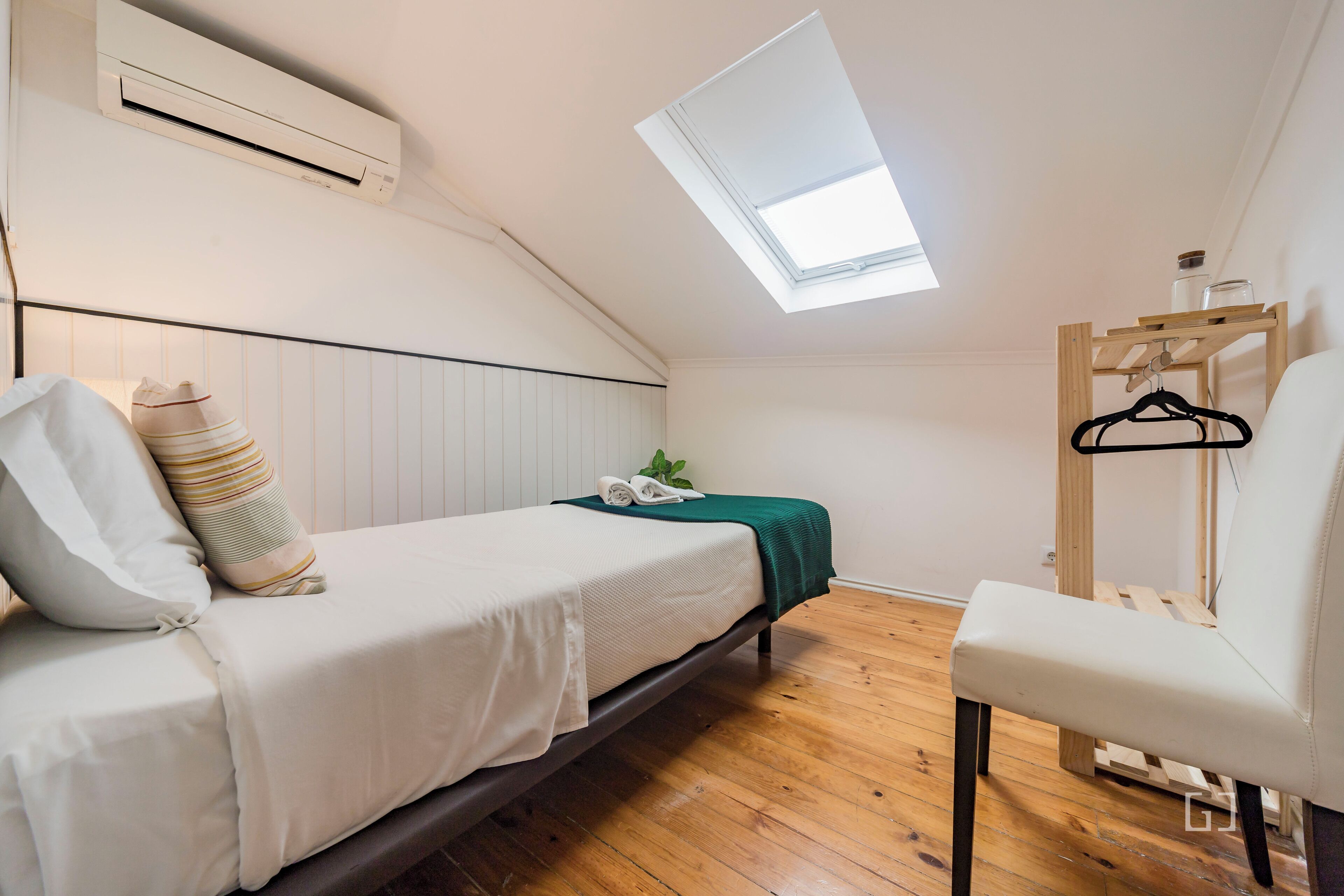 Una habitación pequeña y bien iluminada con techo inclinado, una cama con almohadas, una silla y un estante de madera con un objeto colgando.