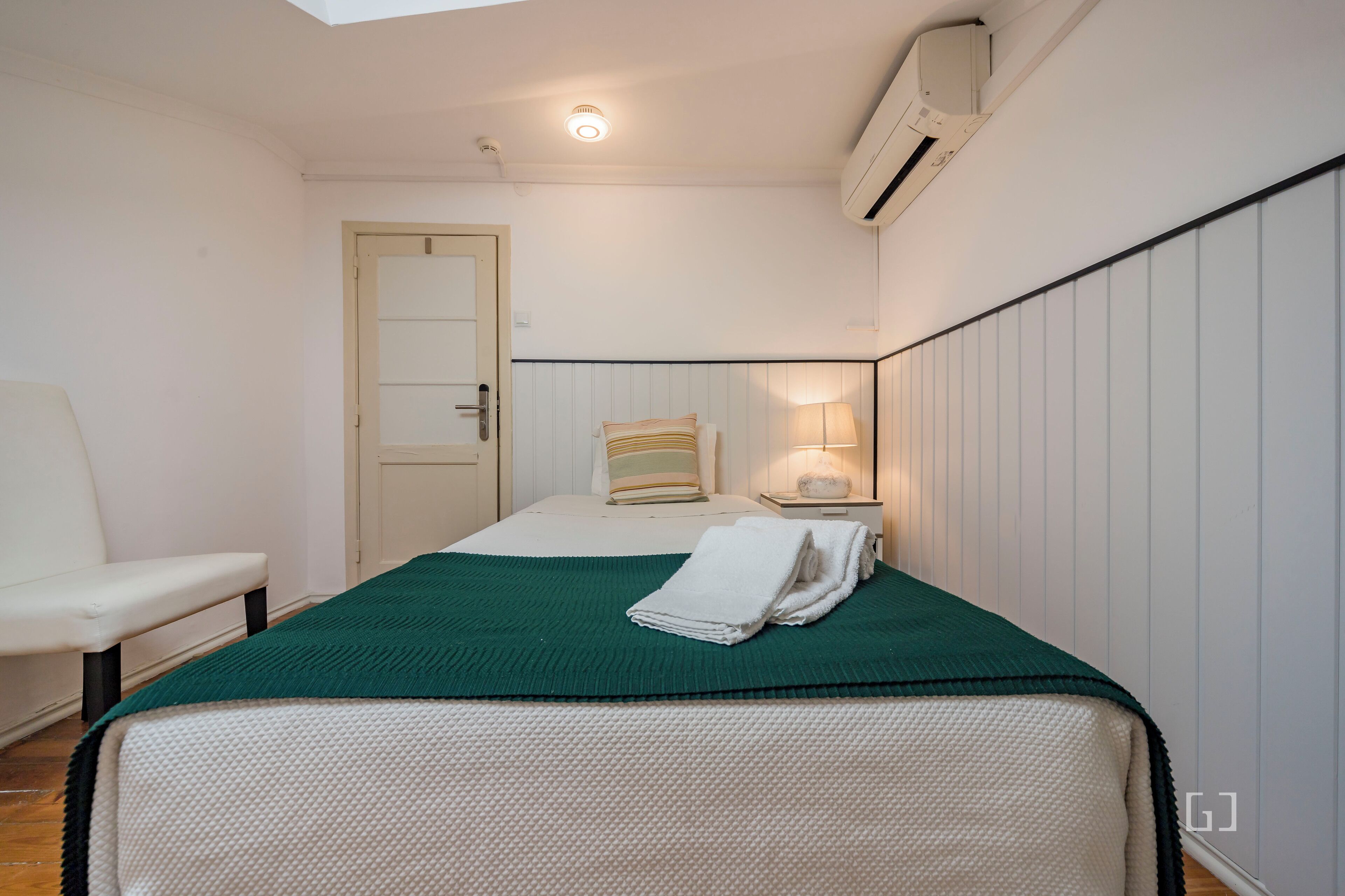 A neatly arranged bedroom with a bed, a chair, a bedside table with a lamp, and an air conditioning unit on the wall.