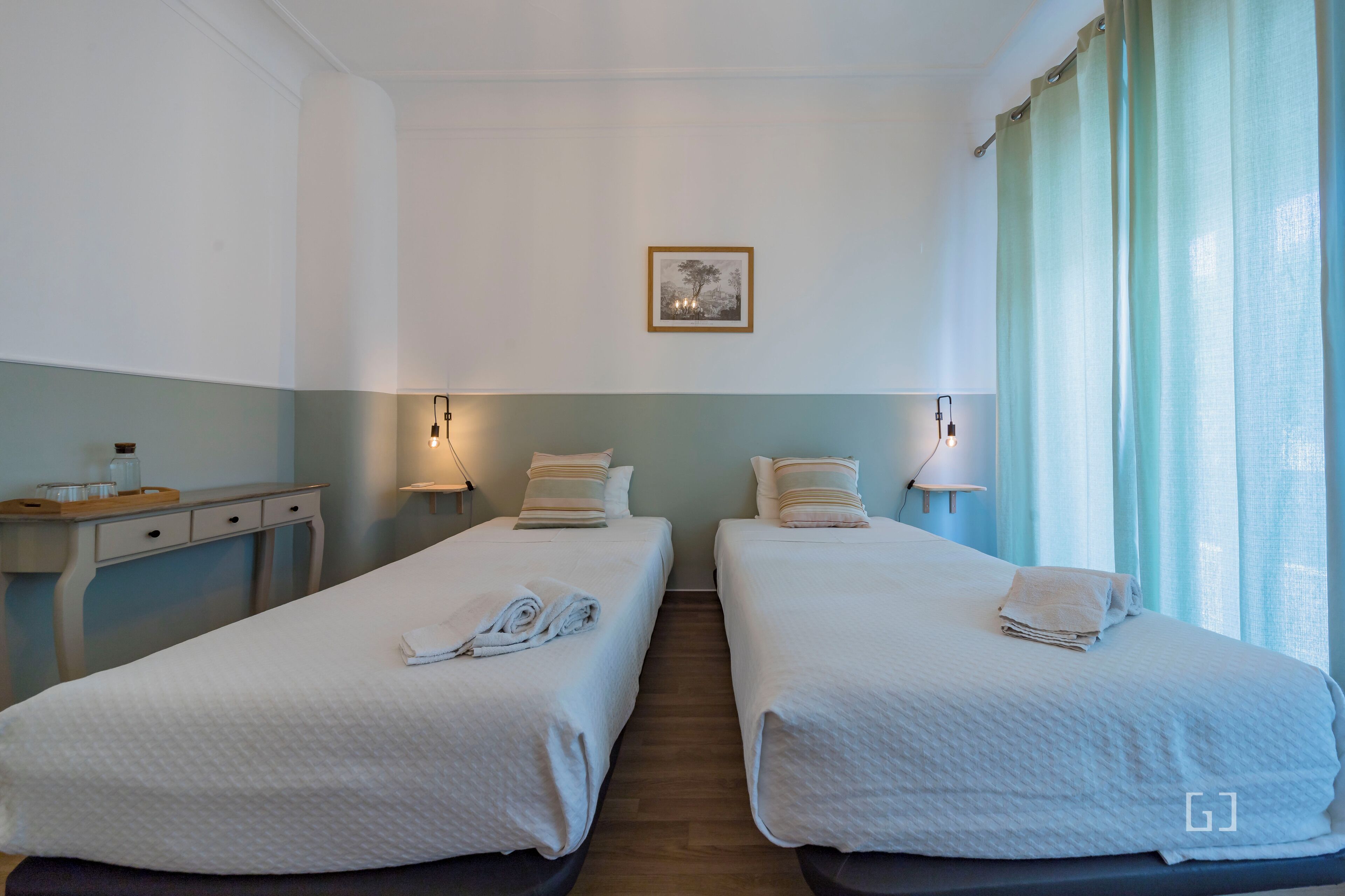 Habitación de hotel con dos camas individuales, un escritorio, una silla, una mesita de noche con una lámpara y un cuadro en la pared.
