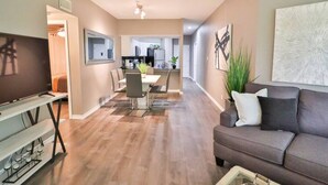 A modern living room with a flat-screen TV, a gray sofa, and a dining area with a table and chairs.