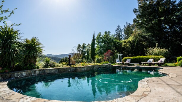 A swimming pool with a stone border, surrounded by greenery and a view of hills. - Farmhouse on Vineyard (Redwood Valley)