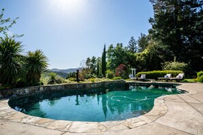 A swimming pool with a stone border, surrounded by greenery and a view of hills. - Farmhouse on Vineyard (Redwood Valley)