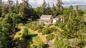 A two-story house surrounded by dense forest, with a visible patio area and a clear sky. - Farmhouse on Vineyard (Redwood Valley)