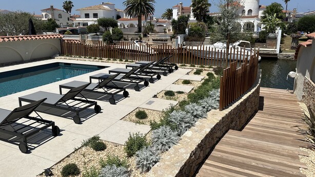 A pool area with lounge chairs, a wooden deck, and a view of buildings in the background. - Beautiful villa on wide canal with 15m mooring and pool! (Empuriabrava)