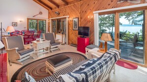 A cozy cabin interior with wooden walls and ceiling, a dining area with red chairs, a sofa with a patterned throw, and a television.