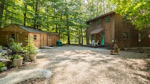 A rustic cabin with a gravel driveway, surrounded by dense forest.
