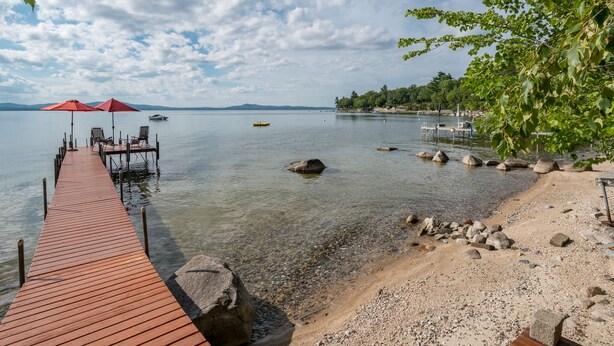 A wooden pier extending into a calm lake with clear water, surrounded by pebbles and rocks.