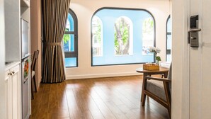 Quarto com piso de madeira, uma janela grande com vista para árvores, cortina e uma cadeira.