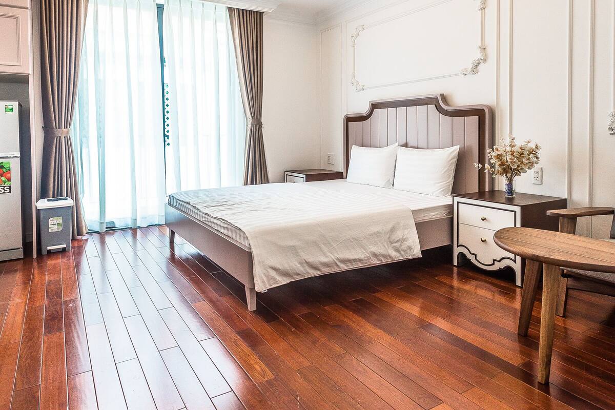 Une chambre d’hôtel avec un plancher en bois, un lit avec du linge de lit blanc, une table de chevet, une table et un plafond doté d’un éclairage encastré.