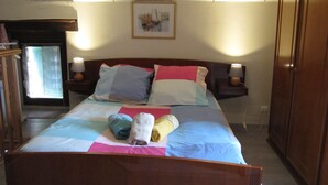 Un dormitorio con cama, almohadas, una mesita de noche, una lámpara, una ventana con cortinas, un cuadro en la pared y un armario.