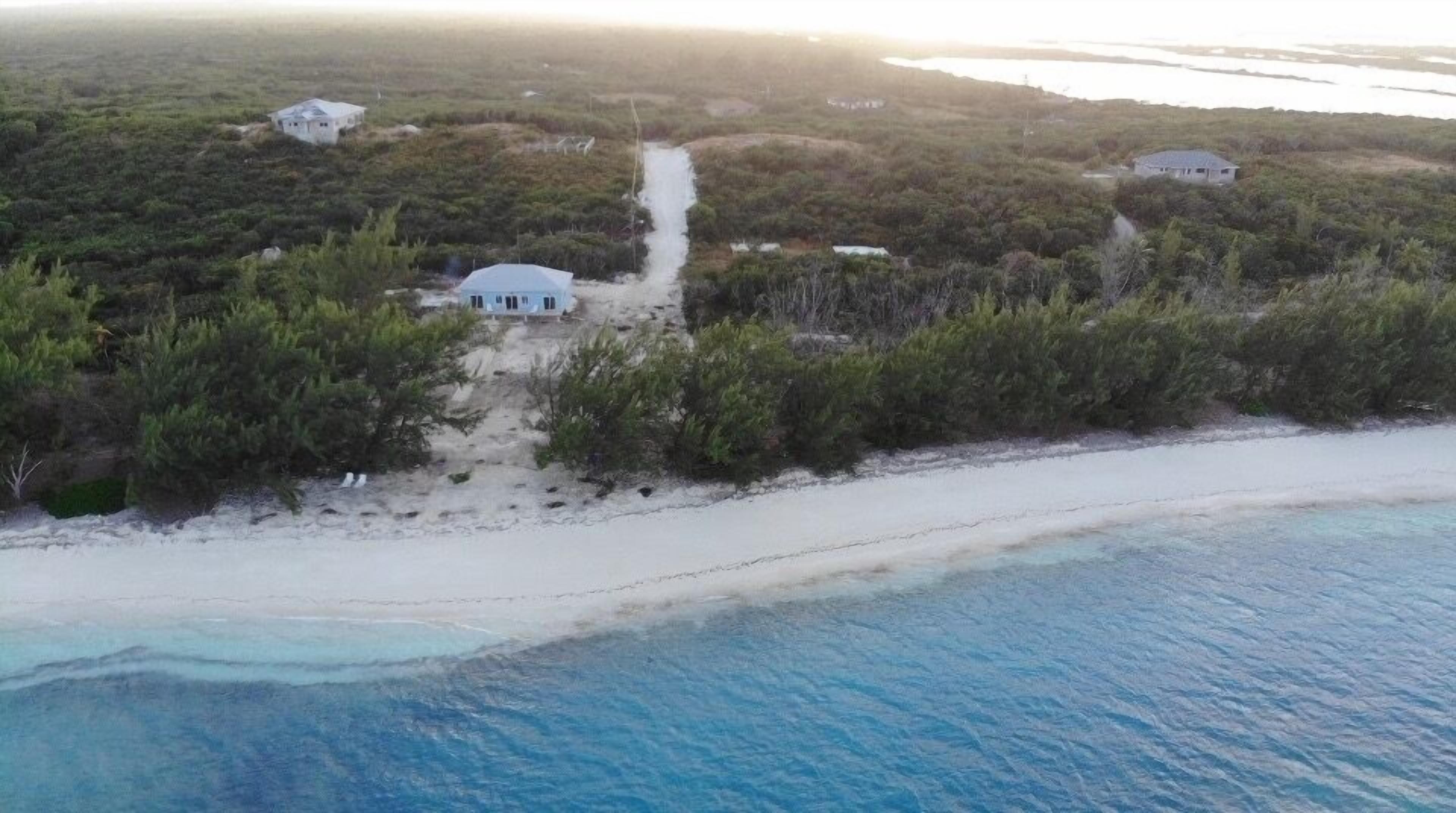 An aerial view of a coastal property with a sandy beach, a clear blue ocean, and a few buildings surrounded by dense vegetation.