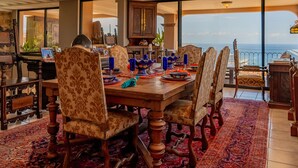 Comedor con una mesa de madera dispuesta para seis personas, rodeado de sillas con estampado, y vistas al océano a través de amplias ventanas.