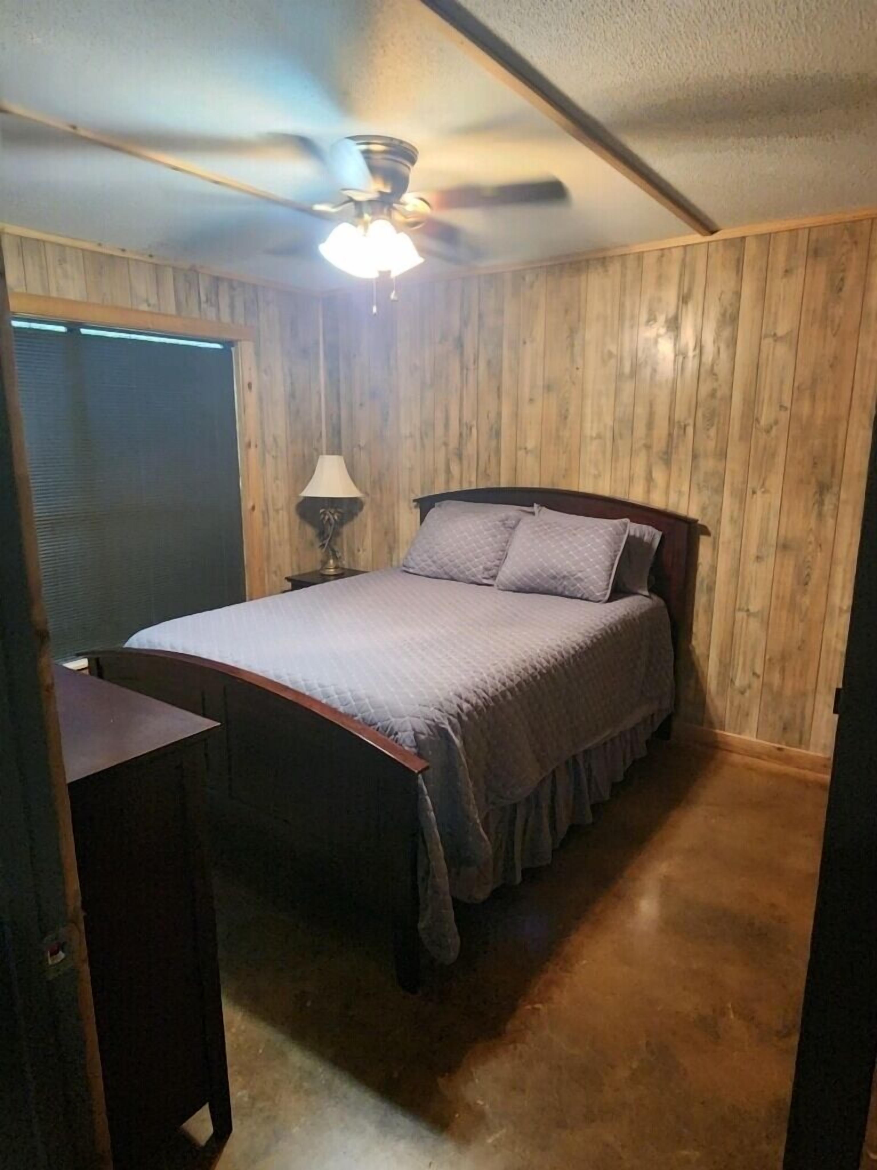 Makuuhuoneessa on puupaneloitu seinä, sängyssä on pääty, yöpöydällä on lamppu ja katossa on kattotuuletin.