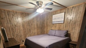 Une chambre avec un lambris en bois, un lit recouvert d’une courtepointe bleue, un ventilateur de plafond et un panneau décoratif encadré sur lequel on peut lire « MAISON AU BORD DU LAC ».