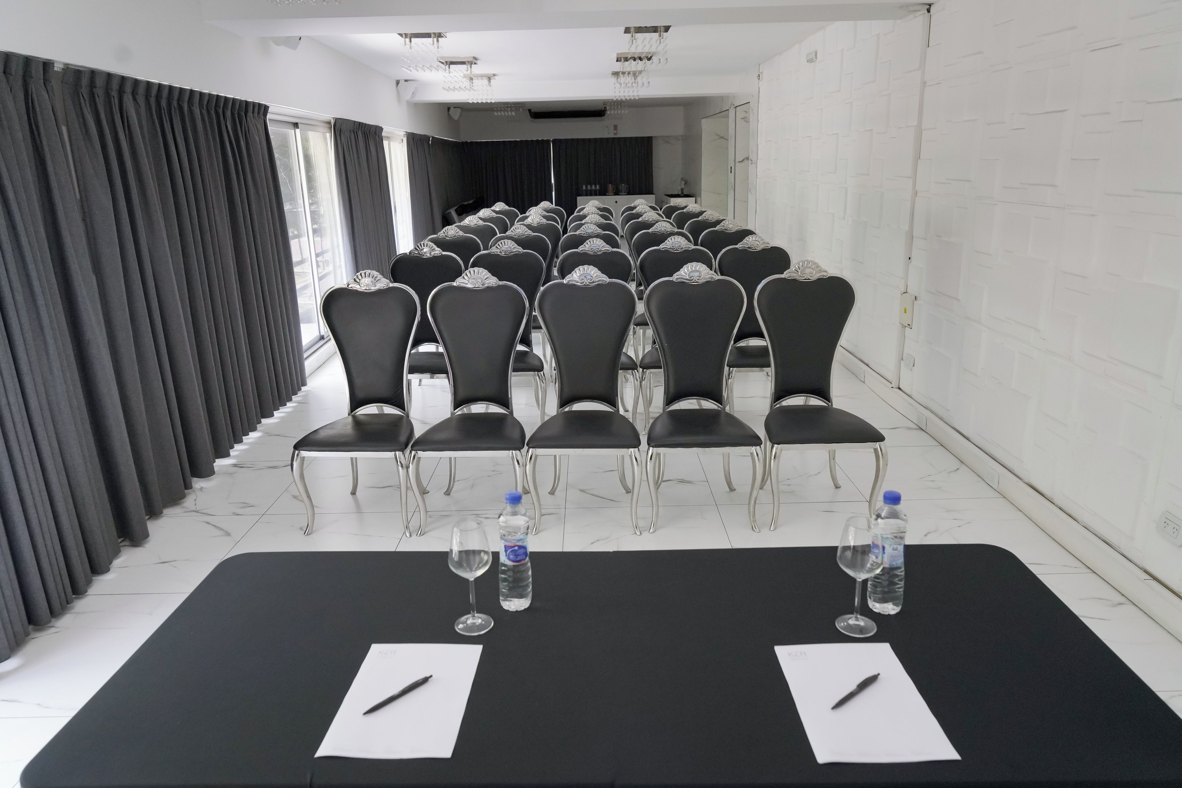 Un salón de conferencias con filas de sillas, una mesa larga con botellas de agua y vasos, y un piso cubierto de baldosas blancas.