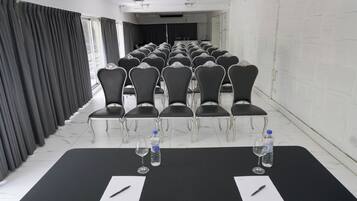 Un salón de conferencias con filas de sillas, una mesa larga con botellas de agua y vasos, y un piso cubierto de baldosas blancas.