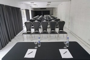 Un salón de conferencias con filas de sillas, una mesa larga con botellas de agua y vasos, y un piso cubierto de baldosas blancas.