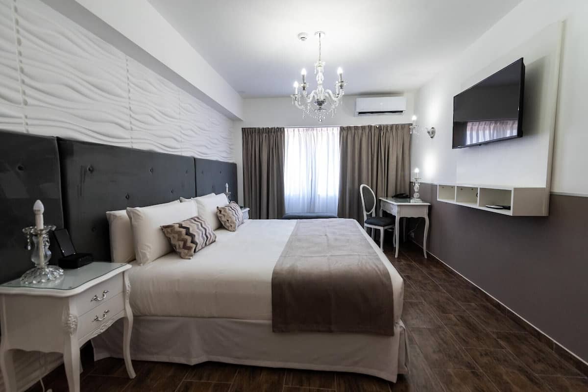 A modern hotel room with a large bed, a desk, a chair, a TV, and a chandelier.