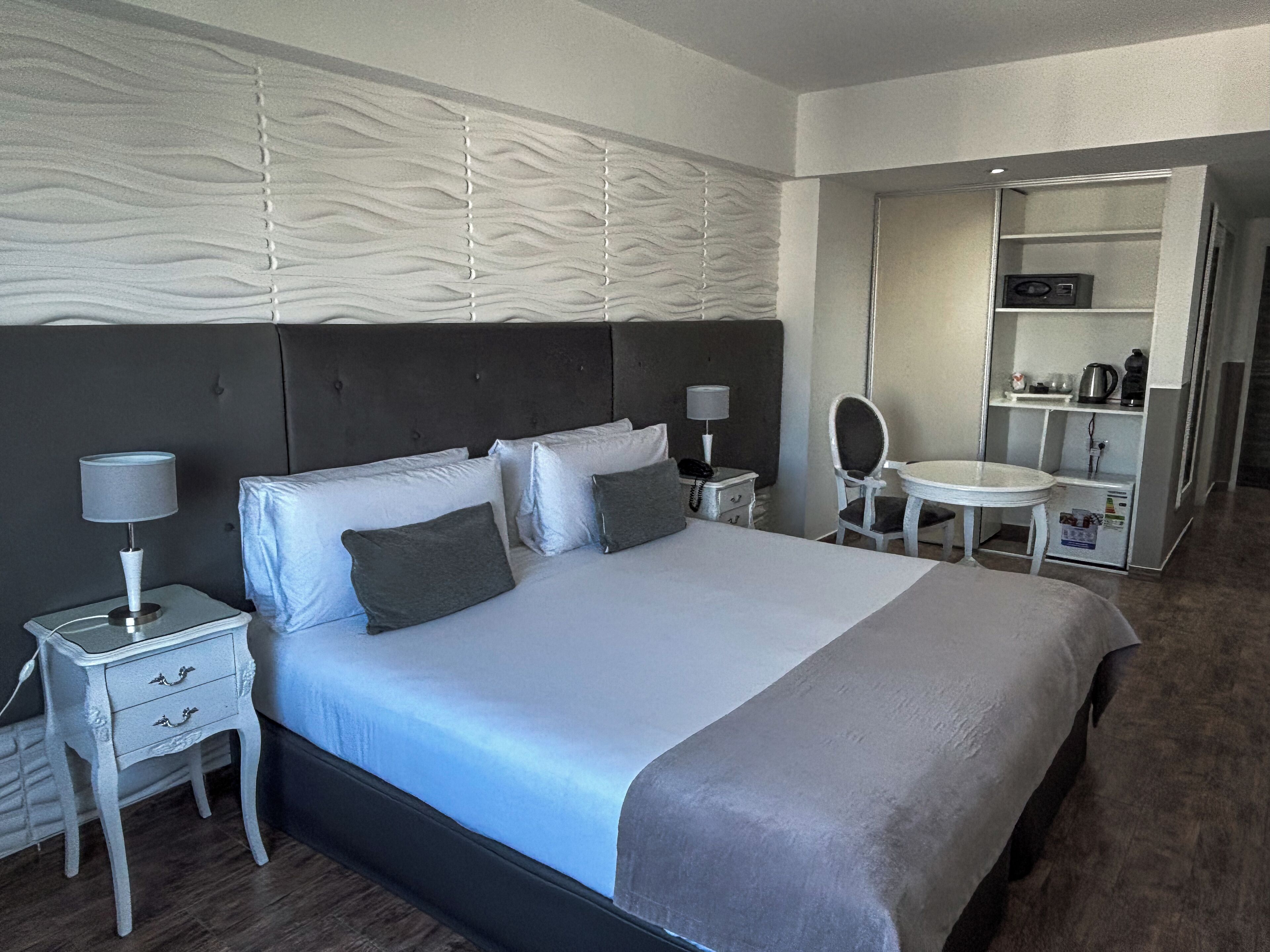 A hotel room with a bed, bedside tables, a mirror, a small table, and a kitchenette area.