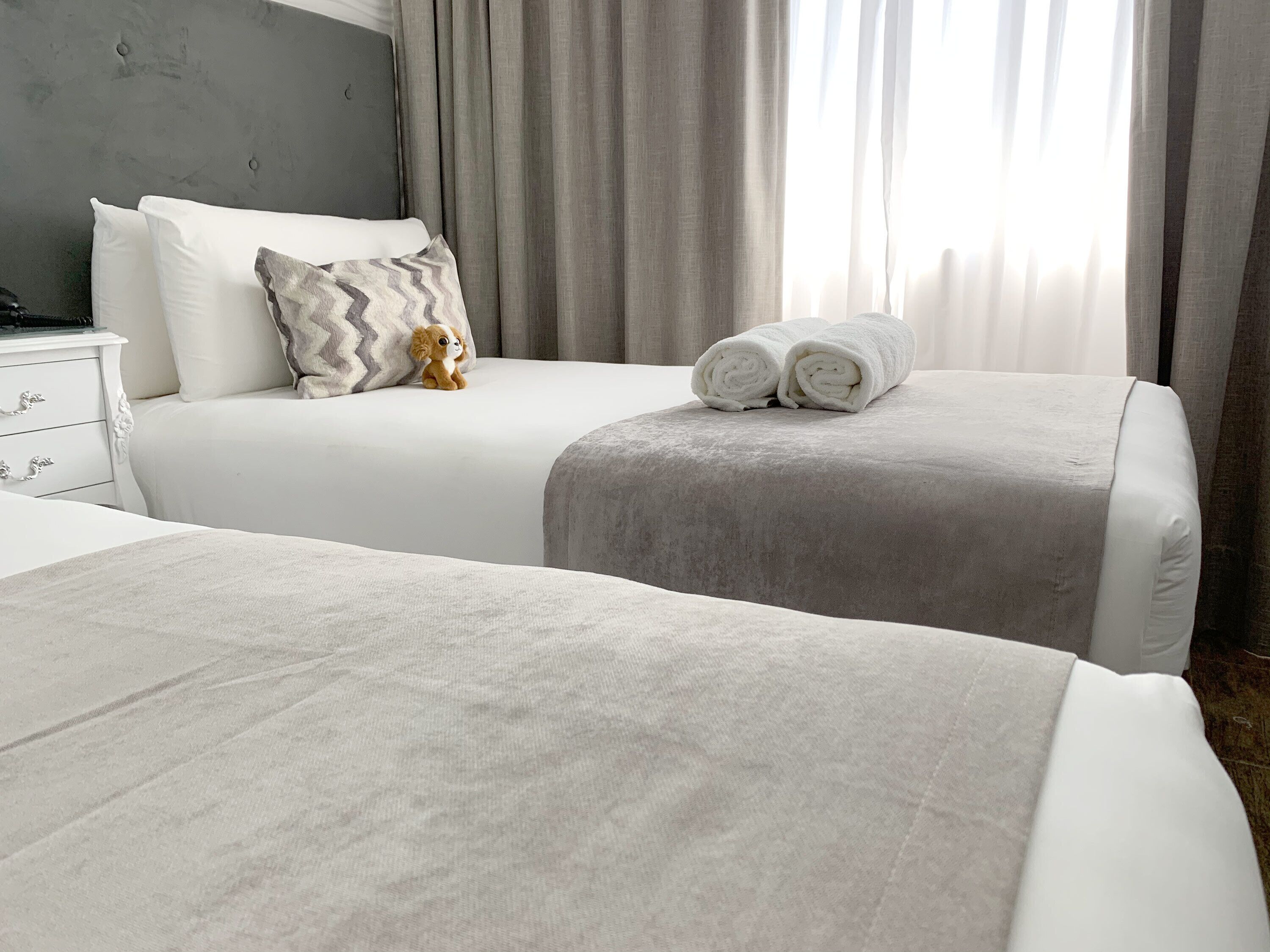 Habitación de hotel con dos camas, una mesita de noche y un cabecero gris.