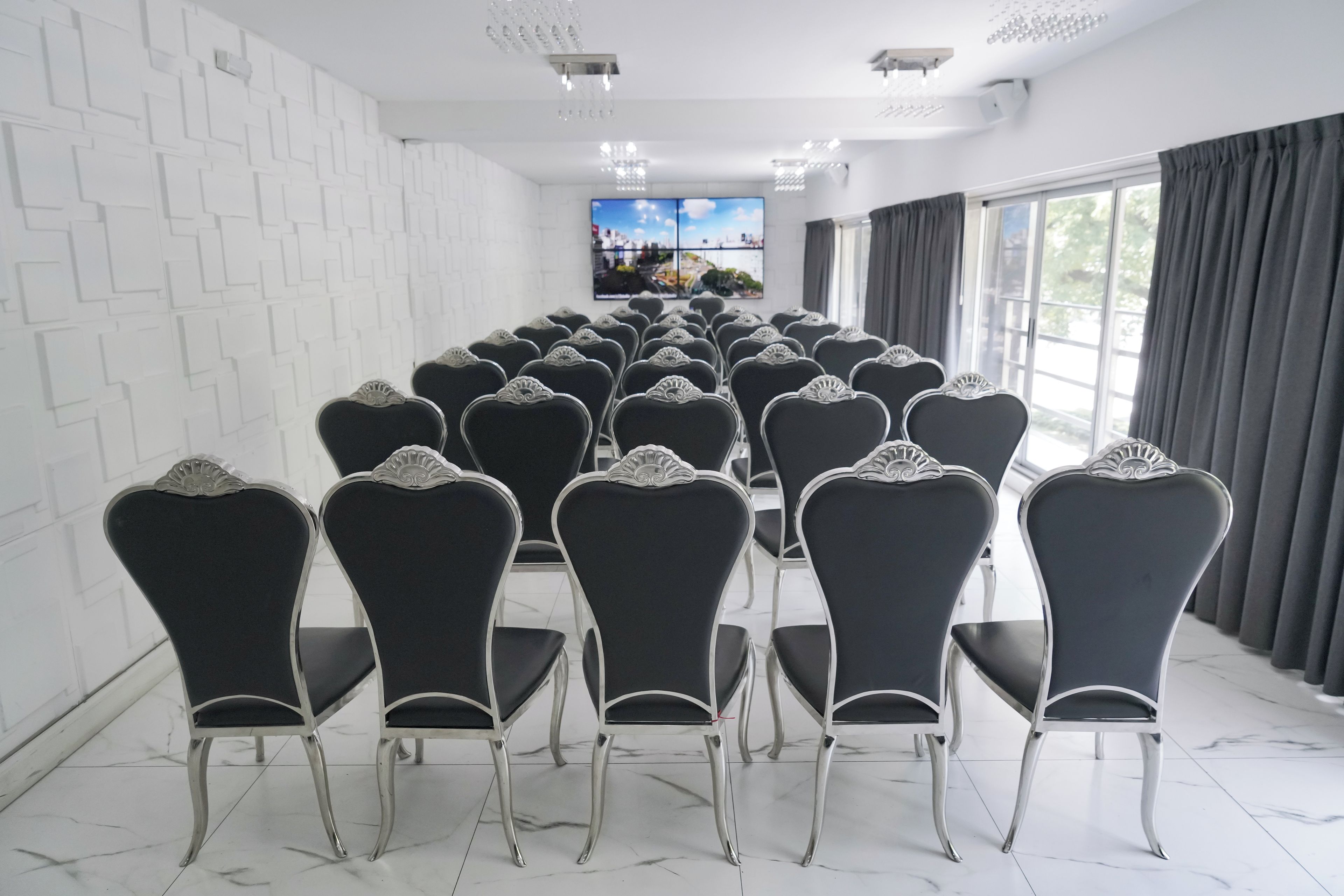 Un salón de conferencias con filas de sillas orientadas hacia una pantalla grande, paredes blancas y un ventanal con cortinas.