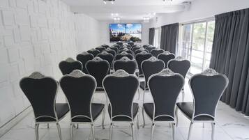 Un salón de conferencias con filas de sillas orientadas hacia una pantalla grande, paredes blancas y un ventanal con cortinas.