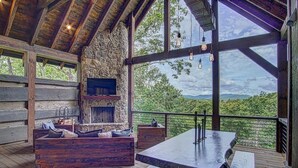 A rustic cabin with a stone fireplace, wooden furniture, and a large window overlooking a forested landscape. - Luxury Cabin with amazing sunsets! Close to Downtown Blue Ridge! (Blue Ridge)