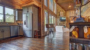 A rustic kitchen with a large refrigerator, wooden cabinets, and a stone wall. There are two bar stools and a television mounted on the wall. - Luxury Cabin with amazing sunsets! Close to Downtown Blue Ridge! (Blue Ridge)