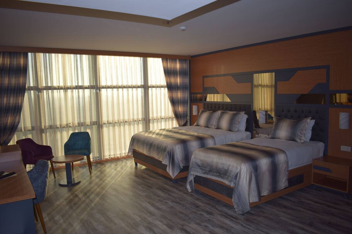 A hotel room with two beds, a desk, a chair, and large windows with curtains.