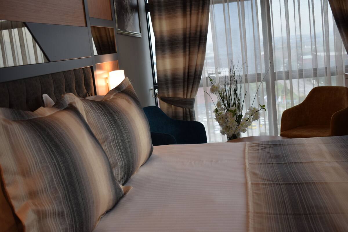 A modern hotel room with a large bed, a view of the city through sheer curtains, and a vase with flowers on the nightstand.