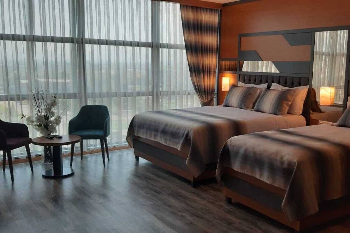 A hotel room with two beds, a sitting area with a table and chairs, and large windows with sheer curtains.